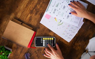 Mão feminina utilizando uma calculadora ao lado de gráficos em papel sobre uma mesa de madeira.