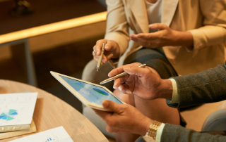 Duas pessoas discutindo gráficos financeiros exibidos em um tablet durante uma reunião. Uma delas segura uma caneta e gesticula, enquanto a outra aponta para o tablet com outra caneta. Há documentos com gráficos em uma mesa de madeira ao lado